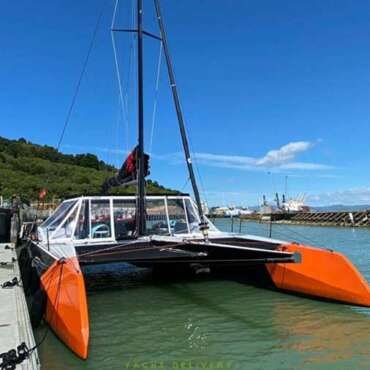 This image shows a multi-hull boat, possibly a catamaran, with a black mast and orange hulls. It's docked in a harbor with a clear blue sky in the background delivered by yacht delivery solutions skipper and crew